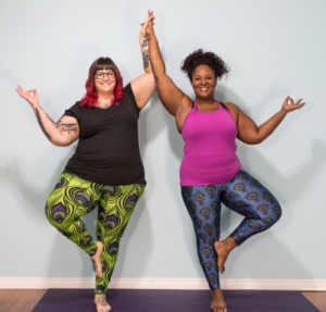 2 larger-bodied women in tree pose, holding hands in the air and smiling at the camera 