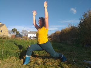 Louise in warrior pose wearing a yellow t-shirt, jean and blue wellies in a field with a blue sky behind.