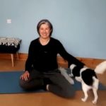 Louise yoga teacher sat on a blue mat smiling at the camera. She's stroking a small, v cute black and white dog