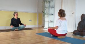 Yoga teacher smiling at student as they both sit on mats in a yoga class