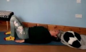 Louise Yoga teacher, lying on her back with knees bent, preparing for 2 foot support pose. A small black and white dog is curled up asleep above Louise's head