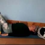 Louise Yoga teacher, lying on her back with knees bent, preparing for 2 foot support pose. A small black and white dog is curled up asleep above Louise's head