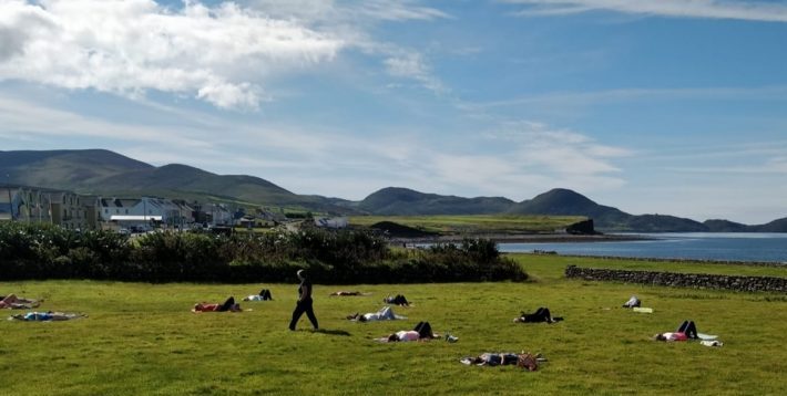 Several people lying on the grass in a relaxing yoga pose while the teacher walks among them. In the background are the green hills and blue sea of Waterville in County Kerry