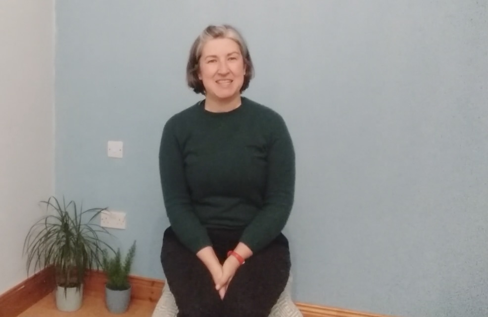 Louise the yoga teacher sat on a stool in a dark green jumper with her hands in her lap