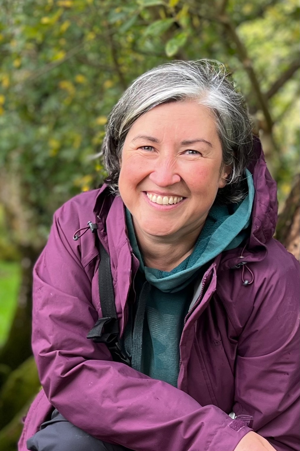 Louise smiling at camera in front of trees. She has greying dark hair and is wearing a purple raincoat