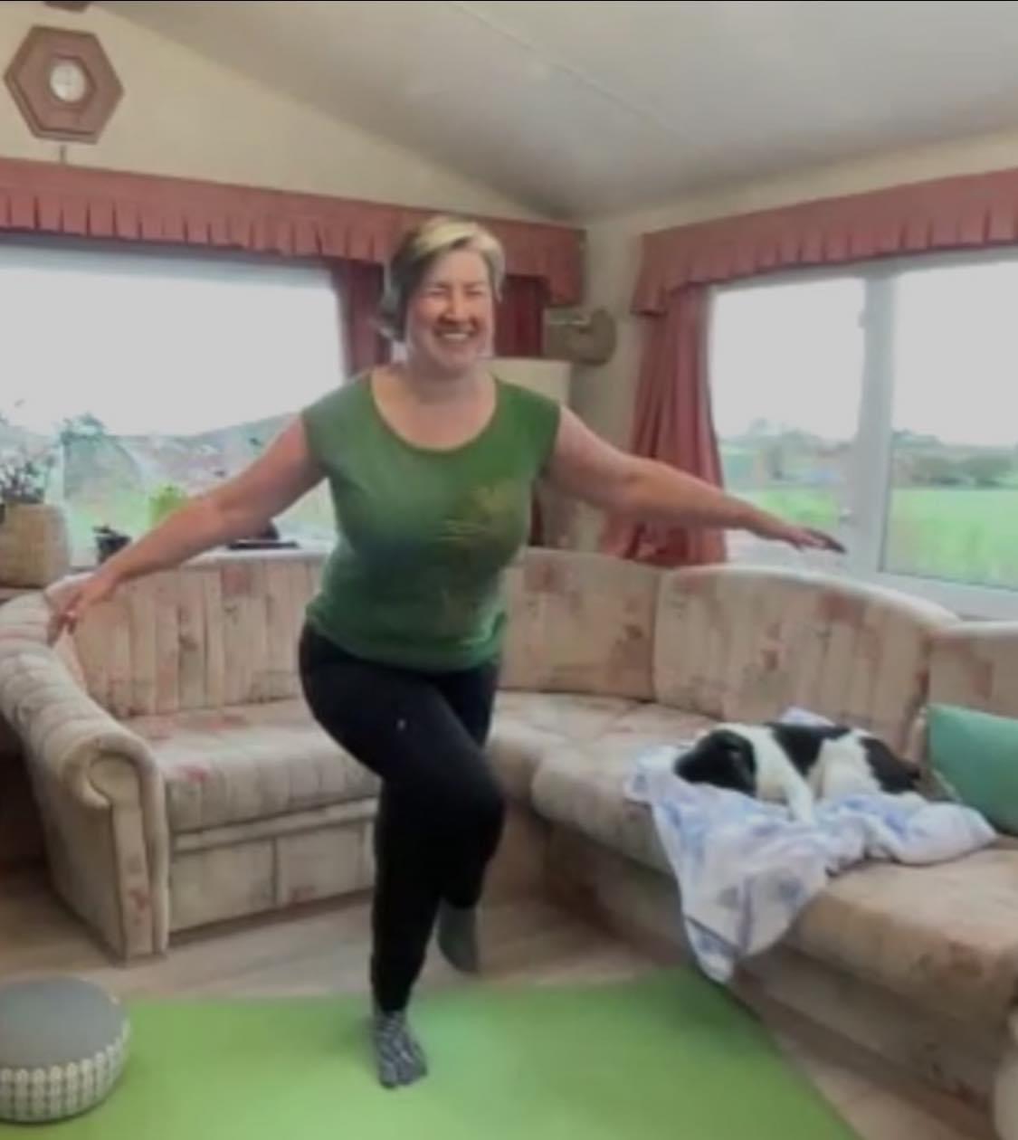 Louise standing on one leg with the other crossed over in eagle pos. Her arms are outstretched and she's smiling. She's standing on a green yoga mat and Rosie the small black and white dog is sleeping on a blanket on the sofa next to her.