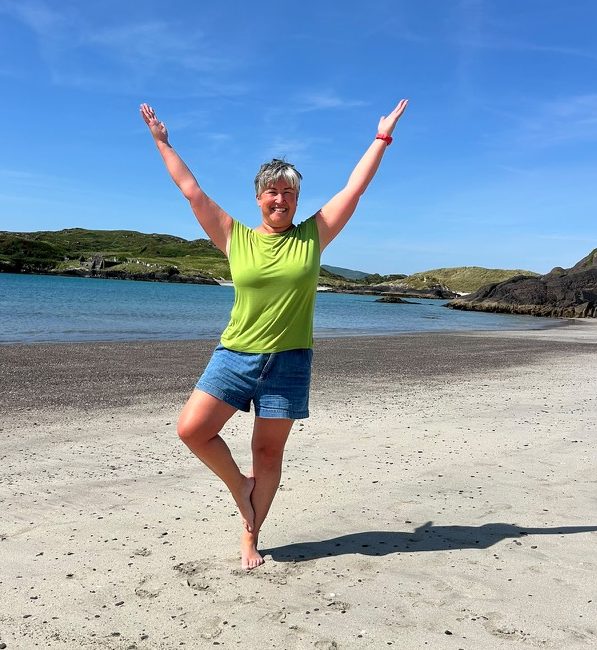 Louise stands in tree pose on the sand with the sea and hills behind her. She's smiling and wears a green t short with blue shorts. The sky is blue and it's very sunny.
